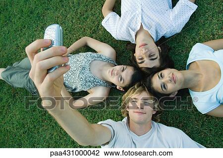 Stock Image - Group of young friends lying on grass, heads together, one young man taking photo with cell phone. Fotosearch