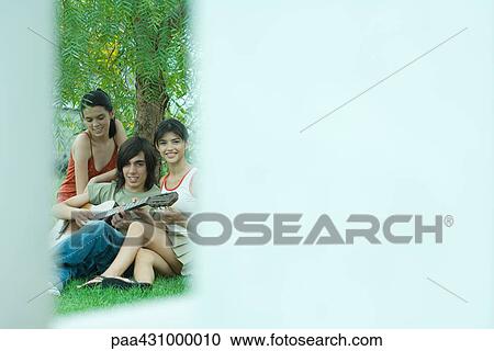Stock Image - Group of young friends sitting by tree while one plays guitar. Fotosearch