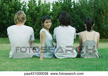 Group of young friends sitting on grass, rear view, one young woman looking over shoulder at camera View Large Photo Image Stock Photo - Group of young friends sitting on grass, rear view, one young woman looking over shoulder at camera. Fotosearch