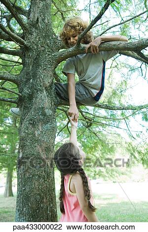 Stock Image - Boy sitting in tree, reaching down to hold girl's hand. Fotosearch