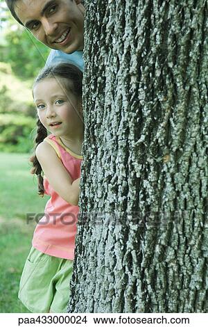 Picture - Girl and father peeking around tree trunk. Fotosearch