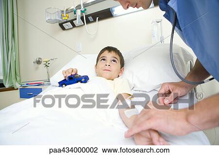 Boy lying in hospital bed holding toy car while intern checks pulse View Large Photo Image Stock Image - Boy lying in hospital bed holding toy car while intern checks pulse. Fotosearch