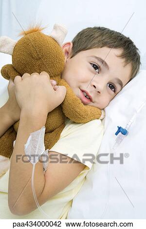 Stock Image - Boy lying in hospital bed with IV drip in arm, hugging stuffed animal. Fotosearch