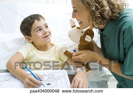 Boy sitting in hospital bed coloring while woman holds up stuffed toy View Large Photo Image Stock Photo - Boy sitting in hospital bed coloring while woman holds up stuffed toy. Fotosearch