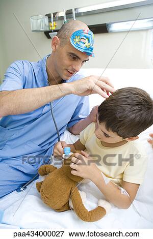 Intern sitting next to boy in hospital bed, looking into boy's ear while boy imitates him with stuffed animal View Large Photo Image Stock Photo - Intern sitting next to boy in hospital bed, looking into boy's ear while boy imitates him with stuffed animal. Fotosearch