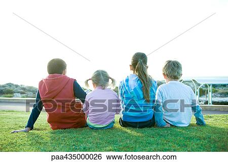 Stock Photograph - Four children sitting on grass, rear view. Fotosearch