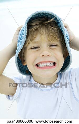 Stock Photograph - Boy holding hat and smiling on beach, portrait. Fotosearch