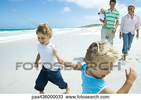 Stock Image - Brother and sister running and laughing on beach in front of parents with baby. Fotosearch