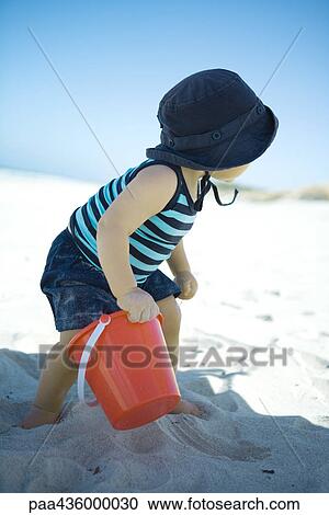 Stock Image - Child standing on beach holding sand bucket. Fotosearch
