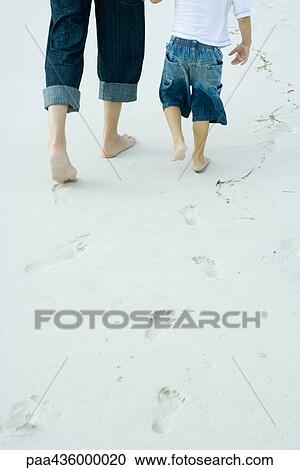 Stock Image - Father and son walking on sand, low section, rear view. Fotosearch
