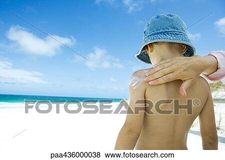 Stock Photo - Female hand applying sunscreen to boy's bare back on beach. Fotosearch