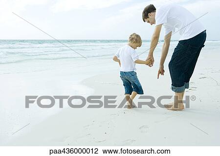 Stock Image - Man and son on beach, man pointing to sand. Fotosearch