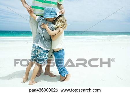Stock Photography - Man standing on beach with son and daughter hugging his legs, waist down. Fotosearch