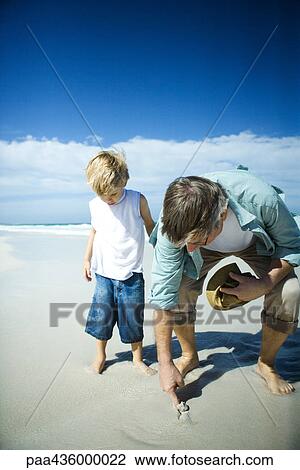 Stock Image - Senior man and grandson on beach, man sticking finger into sand. Fotosearch