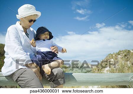 Stock Image - Senior woman sitting on wooden railing, holding baby, dunes in background. Fotosearch
