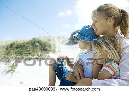 Stock Photo - Woman holding son and daughter on lap, looking toward distance, dune in background. Fotosearch
