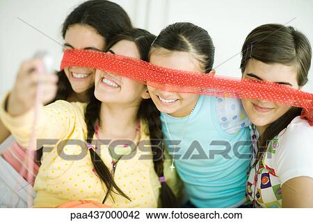 Stock Image - Four young female friends holding scarf in front of eyes, one taking photo with cell phone. Fotosearch