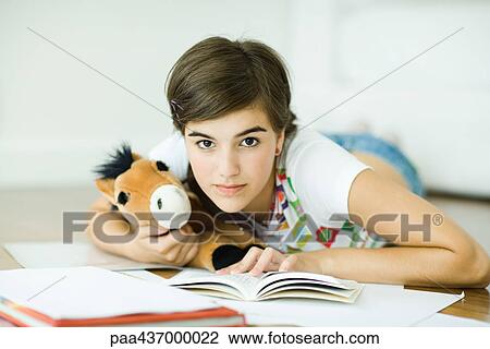 Stock Image - Teen girl lying on floor, doing homework, holding stuffed animal, looking at camera. Fotosearch