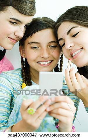 Stock Photo - Three young female friends looking at selves in hand mirror, one holding lipgloss. Fotosearch