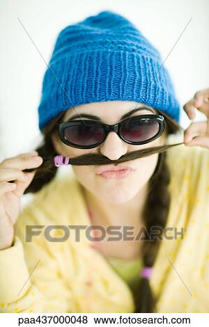 Stock Photo - Young woman wearing knit hat and sunglasses, holding end of pigtail over upper lip, portrait. Fotosearch