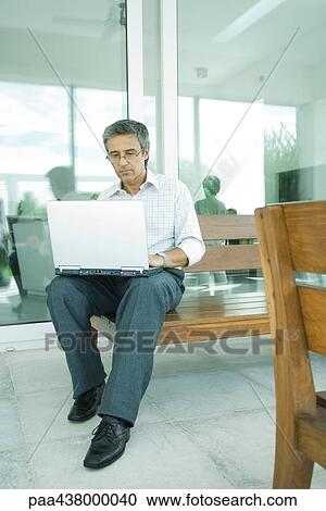 Stock Image - Man sitting, using laptop on patio, full length. Fotosearch