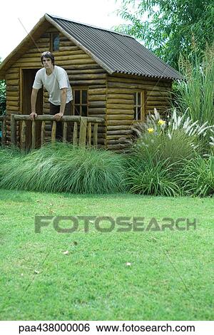 Stock Photograph - Man standing on porch of log cabin, full length. Fotosearch
