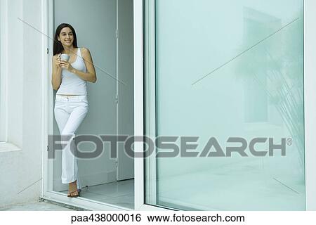 Stock Photograph - Woman standing in doorway with cup, smiling, full length. Fotosearch