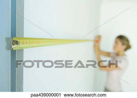 Stock Image - Woman measuring wall with measuring tape, focus on foreground. Fotosearch