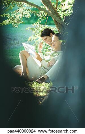 Couple sitting on grass, leaning against tree, reading newspaper together View Large Photo Image Picture - Couple sitting on grass, leaning against tree, reading newspaper together. Fotosearch