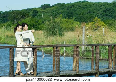 Stock Image - Couple standing on footbridge, woman pointing to the distance. Fotosearch