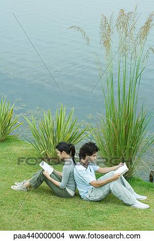 Picture - Young adult couple reading outdoors, sitting back to back on grass by edge of lake. Fotosearch