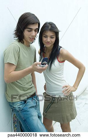 Stock Photo - Young couple leaning against wall, looking at cell phone. Fotosearch