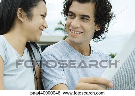 Stock Photograph - Young couple reading newspaper together outdoors, smiling at each other. Fotosearch
