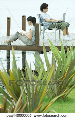 Stock Photography - Young man and woman sitting on dock, reading and using laptop. Fotosearch