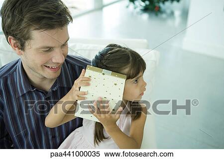 Stock Photography - Girl holding up present to ear while father watches. Fotosearch