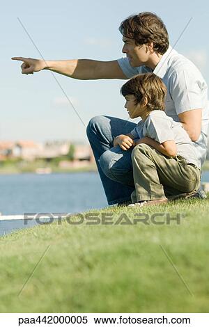 Stock Photography - Father and son sitting by edge of water, looking toward distance. Fotosearch
