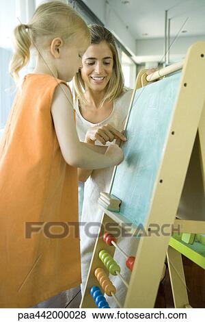 Stock Photo - Woman next to little girl drawing on chalkboard. Fotosearch