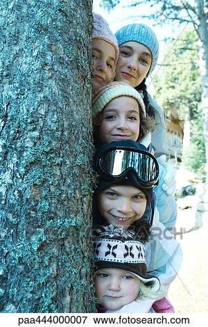 Stock Photo - Kids and teens peeking from behind tree in ski clothes, portrait. Fotosearch