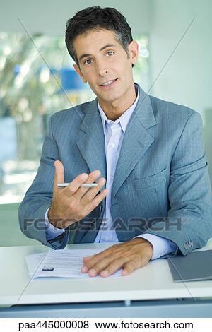 Businessman sitting at table with document, holding pen and gesturing View Large Photo Image Stock Photo - Businessman sitting at table with document, holding pen and gesturing. Fotosearch