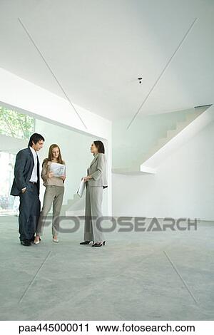 Stock Image - Female real estate agent standing in empty home interior with young couple, full length. Fotosearch