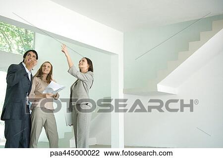 Stock Image - Female real estate agent standing in empty home interior with young couple, full length. Fotosearch
