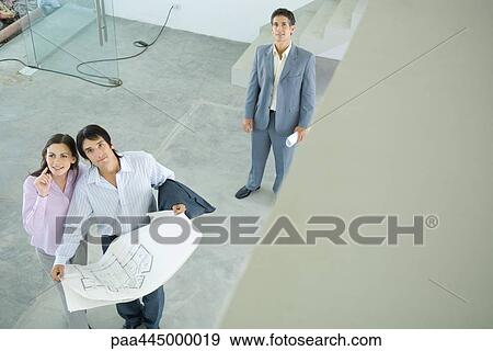 Stock Photo - Male real estate agent standing in home interior with young couple, full length, high angle view. Fotosearch