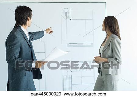 Stock Photo - Man and woman in architect firm, man pointing to blueprints on wall. Fotosearch