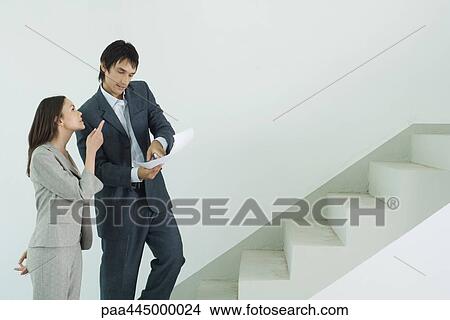 Man and woman in suits standing at foot of stairs, man looking at document, while woman points upwards View Large Photo Image Picture - Man and woman in suits standing at foot of stairs, man looking at document, while woman points upwards. Fotosearch