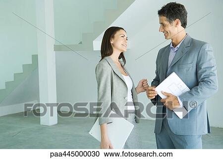 Stock Image - Man and woman in suits standing in bare interior smiling at each other, man holding blueprints. Fotosearch