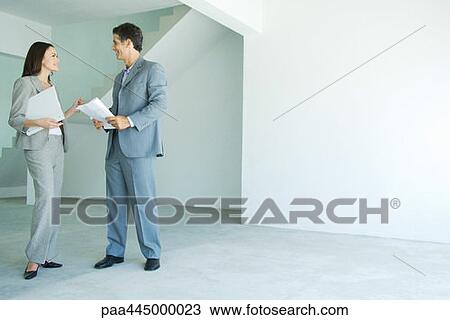 Stock Image - Man and woman in suits standing in empty home interior, smiling, face to face, full length. Fotosearch
