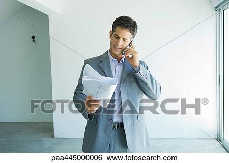 Stock Photograph - Man in suit looking at blueprints, using cell phone. Fotosearch