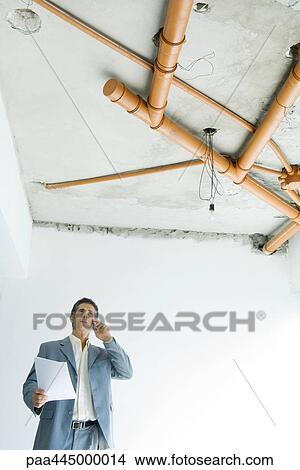 Man in suit standing under bare pipes in home interior, using phone and holding document View Large Photo Image Picture - Man in suit standing under bare pipes in home interior, using phone and holding document. Fotosearch