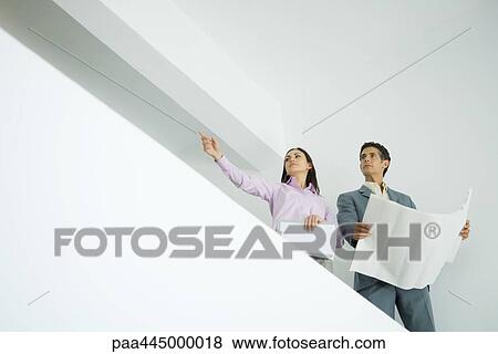 Stock Photo - Woman and man standing with blueprints, woman pointing to distance, low angle view. Fotosearch