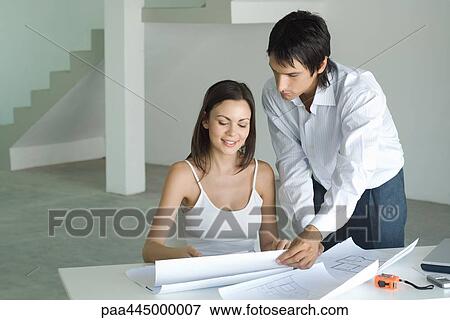 Stock Photo - Woman sitting at table looking at blueprints, man standing, looking over shoulder. Fotosearch
